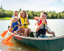 Four people wearing life vests sit in a canoe on a lake, holding paddles with trees visible in the background.