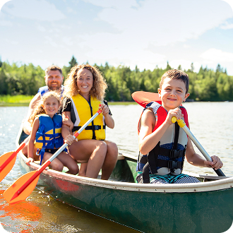 Four people wearing life vests sit in a canoe on a lake, holding paddles with trees visible in the background.