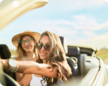 Two people sit in an open‑top car driving on a road in a sunny outdoor setting.