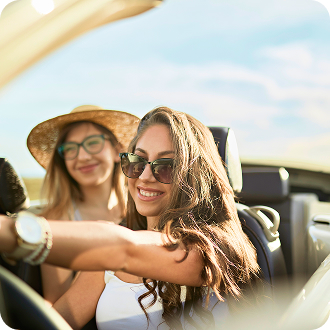 Two people sit in an open‑top car driving on a road in a sunny outdoor setting.