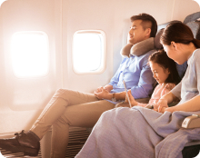 Two adults and a child sit in airplane seats by the window, with one adult wearing a neck pillow and the child using a tablet.