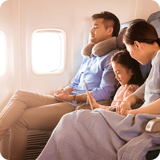 Two adults and a child sit in airplane seats by the window, with one adult wearing a neck pillow and the child using a tablet.
