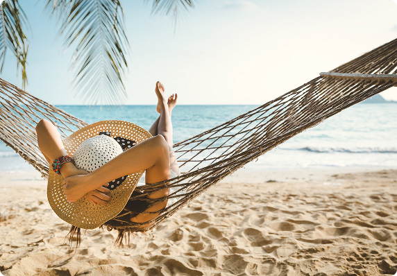 A person lies in a hammock on a sandy beach with the ocean in the background.