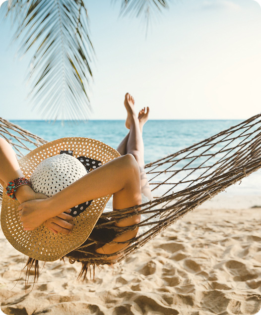 A person lies in a hammock on a sandy beach with the ocean in the background.