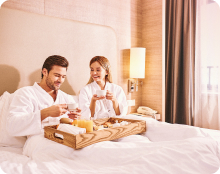 Two people wearing white robes sit on a hotel bed with a breakfast tray holding cups, food, and juice.