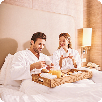 Two people wearing white robes sit on a hotel bed with a breakfast tray holding cups, food, and juice.
